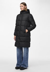 Piumino nero con cappuccio; presenta cuciture orizzontali, tasche laterali e una cerniera frontale. Indossato con jeans in denim a gamba ampia.