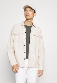 Helebeežikas nööpidega jakk kahe rinnataskuga, kantud tumerohelise henley särgi kohal, valgete pükste ja hele nokamütsiga.