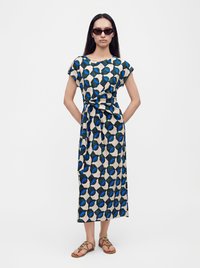 Femme portant une robe bleue et noire à motifs circulaires, des sandales marron et des lunettes de soleil foncées, debout les mains dans le dos.