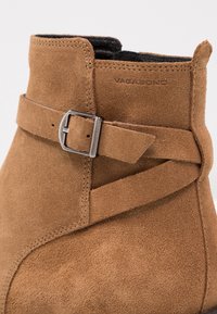 Brun mocka ankelsko med rund tå, prydd med en dekorativ spännslinga och enkel sömnad längs sömmarna. Platt sula.