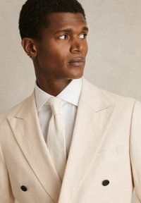 Veste de costume double boutonnage beige clair avec un revers cranté, assortie d'une chemise blanche et d'une cravate claire. Détail de bouton noir sur la veste.