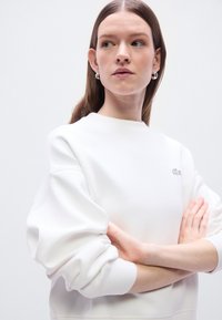 Sweat-shirt blanc à encolure ronde et manches surdimensionnées, arborant un petit logo brodé sur la poitrine. Texture de tissu lisse.