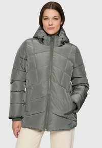 Woman wearing a gray quilted puffer jacket with hood, zipper front, and hands in pockets, standing against a plain light background.