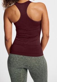 Donna che indossa un top senza maniche color maroon con schiena a corsetta e leggings grigi texturizzati, vista da dietro su uno sfondo semplice.