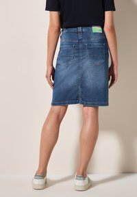 Jupe en denim présentant une coupe A-line classique, de couleur bleu moyen, avec un design à cinq poches et une étiquette au dos. Tissu texturé avec des détails de couture.