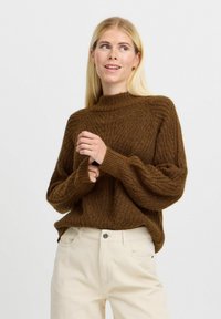 Pulover tricotat maro cu gât înalt, având o textură cu unuțuri și o croială lejeră. Asortat cu pantaloni din catifea ușor colorată.