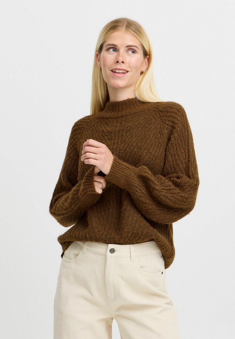 Pulover tricotat maro cu gât înalt, având o textură cu unuțuri și o croială lejeră. Asortat cu pantaloni din catifea ușor colorată.