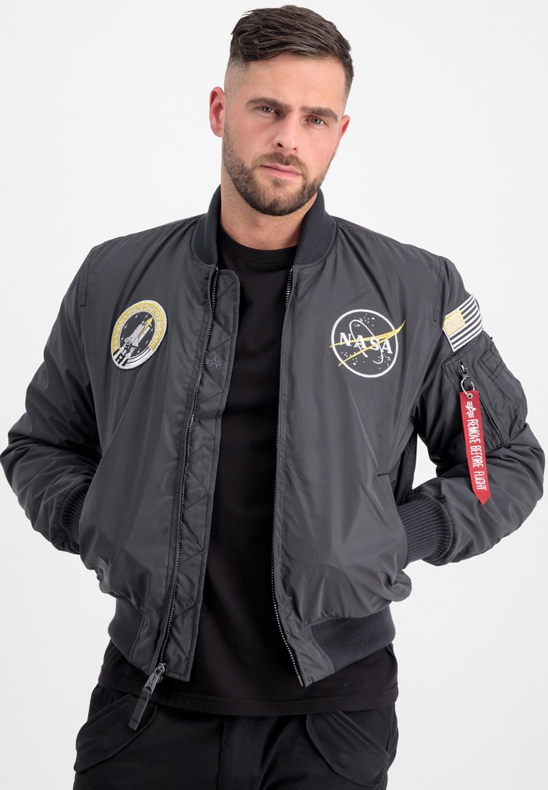 Giacca bomber grigia con emblema NASA, toppe, polsini a coste e etichetta decorativa rossa. Texture liscia e chiusura con zip, con tasche laterali.