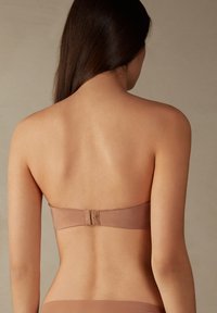 Brązowy biustonosz bez ramiączek z gładkiego materiału, posiadający zapięcie na plecach. Projekt jest minimalistyczny i bezszwowy, podkreślający prostotę.