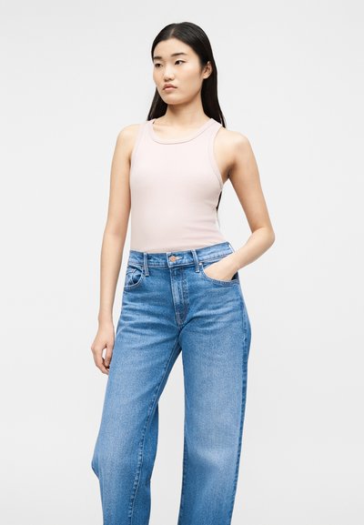Νεαρή γυναίκα με μακριά μαύρα μαλλιά που φοράει αμάνικο ανοιχτό ροζ τοπ και φαρδιά ψηλόμεσα μπλε τζιν, στέκεται με το ένα χέρι στην τσέπη.