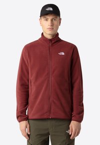 Giacca in pile color bordeaux con colletto alto, chiusura con zip e tasche laterali. Presenta un piccolo logo bianco sul lato sinistro del petto.