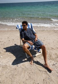 Homme portant des lunettes de soleil, se relaxant pieds nus sur une chaise de plage recouverte d'une serviette rayée, près du rivage océanique sur une plage de sable.