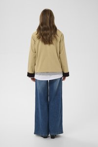 Persona ar gariem brūniem matiem, valkājot bēša krāsas jaku pār pelēkām un baltām drēbēm, platām zilām džinsu biksēm, stāvot ar seju prom uz vienkrāsaina fona.