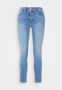 Jeans skinny em denim azul claro, com um corte elevado, cinco bolsos e uma textura ligeiramente desbotada. Fecho clássico com botão e ziper.