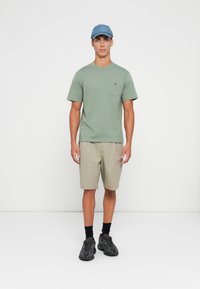Jonge man in een groene T-shirt, beige shorts, zwarte sneakers, zwarte sokken en een blauwe denim pet die tegen een effen witte achtergrond staat.