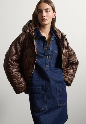 Young woman wears a dark brown quilted hooded jacket over a blue denim dress with large front pockets and button details.