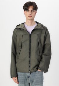 Olivengrønn zip-up jakke med hette, med to frontlommer, teksturert stoff og justerbare mansjetter. Båret over en lys lilla skjorte.