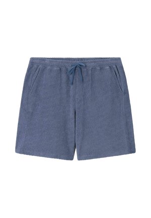 Mann trägt blaues, strukturiertes Poloshirt mit passenden Shorts und beigen Schuhen, hält beige und braune Tasche vor einfarbigem Hintergrund.