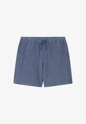 Mann trägt blaues, strukturiertes Poloshirt mit passenden Shorts und beigen Schuhen, hält beige und braune Tasche vor einfarbigem Hintergrund.