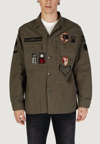 Olijfgroene militaire jas met meerdere patches, twee borstzakken, knoopsluiting en lange mouwen. Heeft een rechte snit en een lichtgewicht stof.