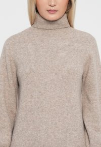 Maglione Beige a collo alto con texture a coste. Materiale morbido e lavorato a maglia, presenta un colletto alto e maniche lunghe.