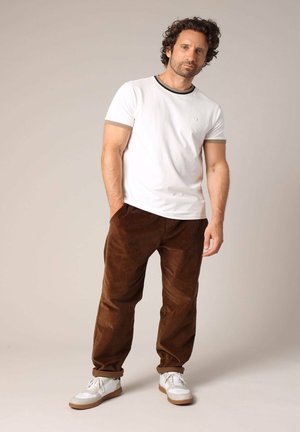 T-shirt blanche à manches courtes avec des bordures sombres, associé à un pantalon en velours côtelé marron et des baskets blanches avec des semelles marron clair.
