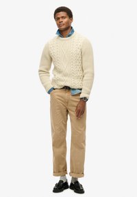 Cremig gestrickter Pullover mit Rautenmuster, darunter ein hellblaues Jeanshemd, beige Cordhosen, schwarze Loafers und eine Armbanduhr.
