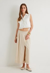 Camicia bianca senza maniche, corta, con due tasche frontali, abbinata a pantaloni beige a gamba larga con tasche laterali profonde e una texture morbida.