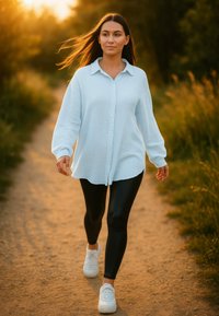 Chemise bleu clair à manches longues, à boutons, avec une coupe décontractée, associée à un legging noir et des baskets blanches, marchant sur un chemin en terre.