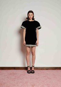 Γυναίκα που φοράει κοντό βελούδινο μαύρο φόρεμα με φουσκωτά μανίκια και βολάν με πουά, στέκεται σε ροζ χαλί μπροστά σε λευκό τοίχο.