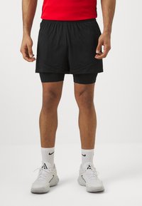Pantalones cortos deportivos negros con un patrón de malla sobre pantalones cortos de compresión negros, usados con zapatillas blancas y calcetines hasta el tobillo con un logo.