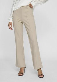 Pantalones de pierna ancha en color beige con una textura suave, que cuentan con costuras verticales. Combinados con sandalias de tacón negro, sobre un fondo claro.