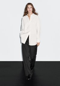 Camicia bianca a righe sottili con vestibilità morbida e chiusura a bottoni, abbinata a pantaloni ampi neri e scarpe nere, su uno sfondo semplice.