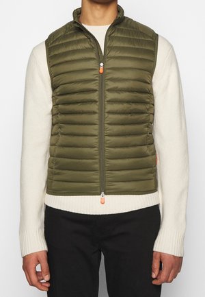 Man die een olijfgroene, gewatteerde mouwloze puffer-jas draagt over een witte geribbelde trui en zwarte broek, staand tegen een effen achtergrond.