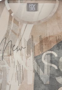Helles beigefarbenes T-Shirt mit abstrakten geometrischen Mustern, versehen mit dunklen Akzenten und einem "Neu"-Druck, der mit metallischen Strasssteinen verziert ist.