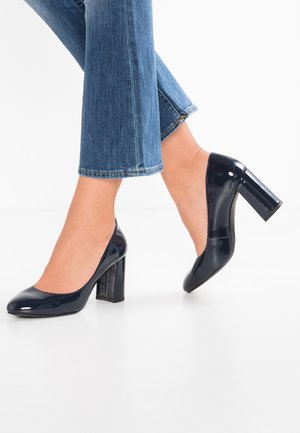 Escarpins en cuir verni marine avec un bout rond et un large talon carré, associés à un jean cropped en denim bleu clair. Texture lisse, design simple.