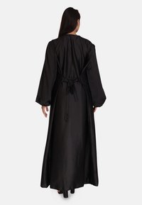 Lang sort satin kjole med brede ærmer, båndlås bagpå og flydende kant, der viser en glat, blank overflade.