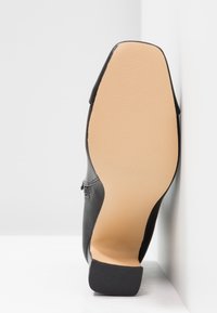 Zapato de cuero negro con parte superior lisa, puntas almendras y suela de goma beige con un patrón de agarre texturizado. Diseño angular y tacón bajo.