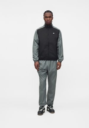 Jeune homme aux cheveux tressés, portant un survêtement noir et gris Nike et des baskets noires Nike, debout devant un fond blanc uni.