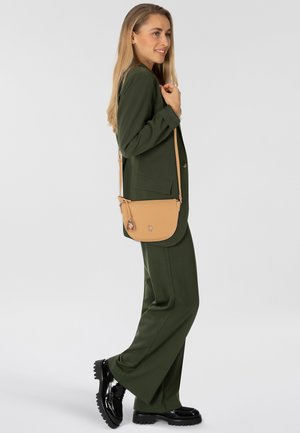Borsa a tracolla marrone con una texture liscia, dotata di chiusura con zip e due charm decorativi. Indossata con un completo verde e scarpe nere.