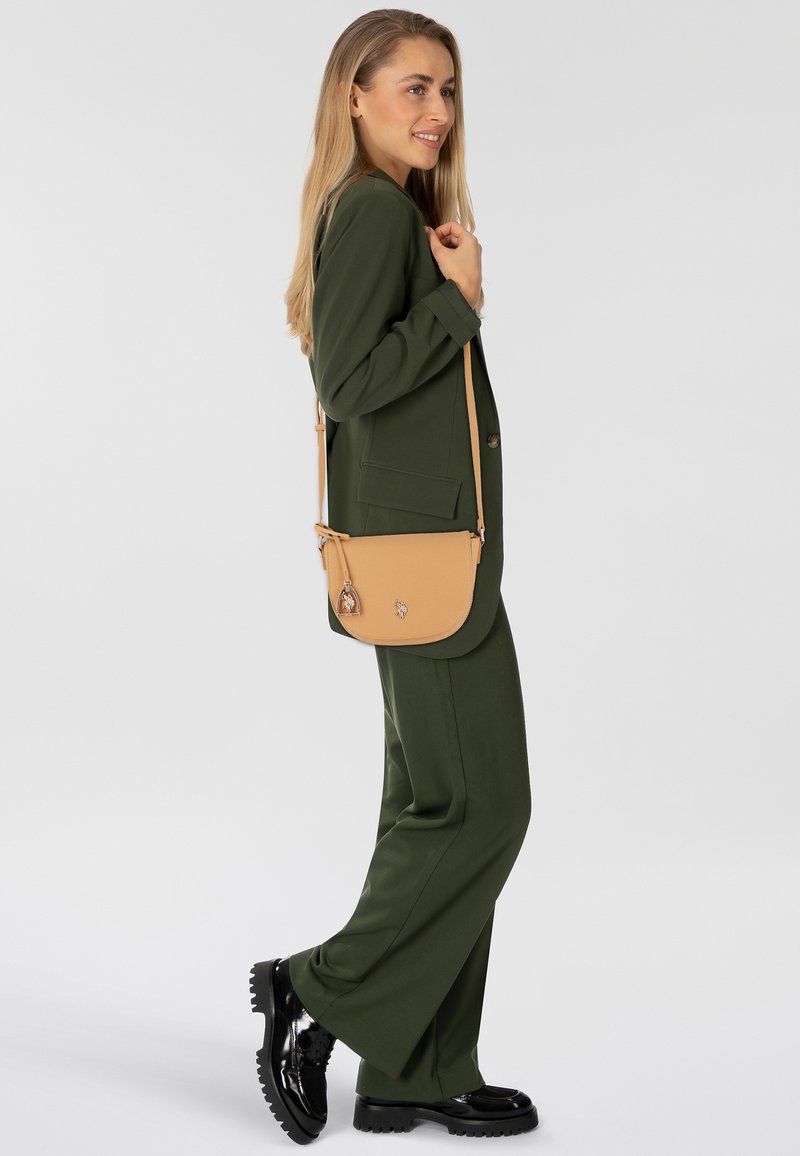 Borsa a tracolla marrone con una texture liscia, dotata di chiusura con zip e due charm decorativi. Indossata con un completo verde e scarpe nere.