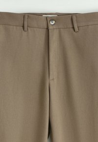 Pantalon beige en tissu lisse et texturé. Comprend une fermeture à bouton, une ceinture standard et des poches latérales.