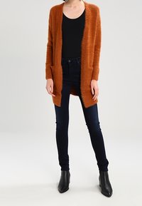 Lång, fluffig cardigan i bränd orange med öppen front och sidofickor, kombinerad med mörkblå skinny jeans och svarta klackade ankelstövlar.