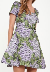 Kortärmad klänning med ett lila blommönster och gröna blad, tillverkad av strukturerad tyg, med en utställd kjoldesign och fyrkantig halsringning.
