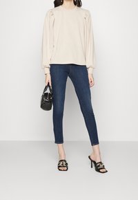 Beige puffärmad topp, mörka tajta jeans, svarta sandaler med guld accenter och en liten svart handväska. Snyggt tyg och skräddarsydd passform.