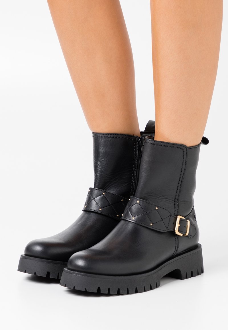 Schwarze Leder-Stiefeletten mit dicken Sohlen, seitlichem Reißverschluss und gestepptem Riemen mit goldener Schnalle, getragen an nackten Beinen.