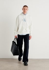 Sudadera blanca con logo azul, superpuesta sobre una camisa azul clara; pantalones amplios negros y zapatos negros; llevando un gran bolso negro.