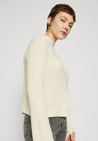 Maglione lavorato a maglia color crema con collo alto, texture a coste e maniche lunghe, indossato con jeans grigi a vita alta.