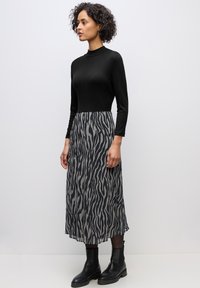 Frau mit lockigem Haar, die ein schwarzes Langarmoberteil, einen grauen und schwarzen Midi-Rock mit Zebramuster, schwarze Strumpfhosen und schwarze Stiefeletten trägt und steht.