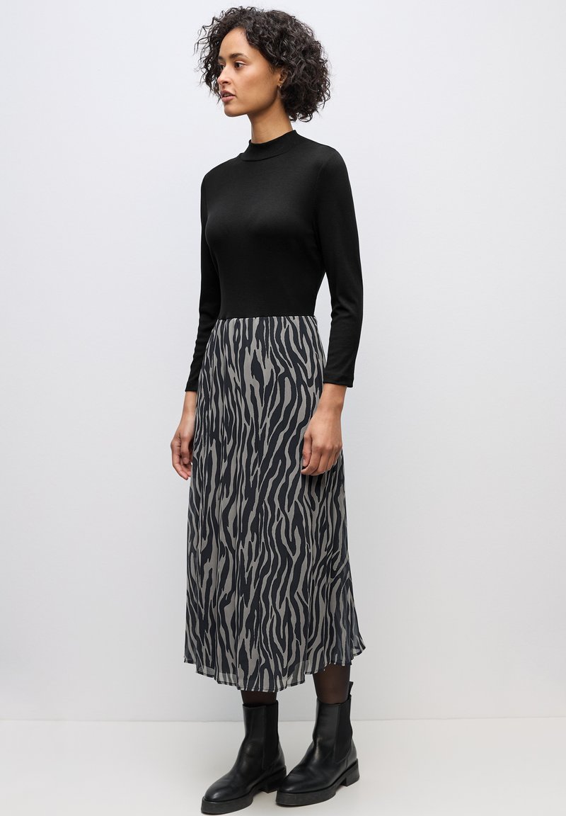 Frau mit lockigem Haar, die ein schwarzes Langarmoberteil, einen grauen und schwarzen Midi-Rock mit Zebramuster, schwarze Strumpfhosen und schwarze Stiefeletten trägt und steht.